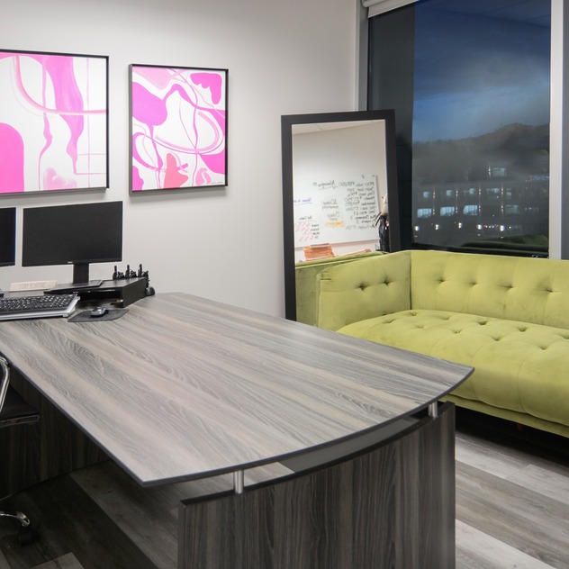 Modern office with a wooden desk, dual monitors, green couch, artwork on the wall, and a window view of a city skyline at night.