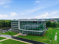 Modern glass office building with green landscaping, cloudy blue sky, and adjacent parking lot.