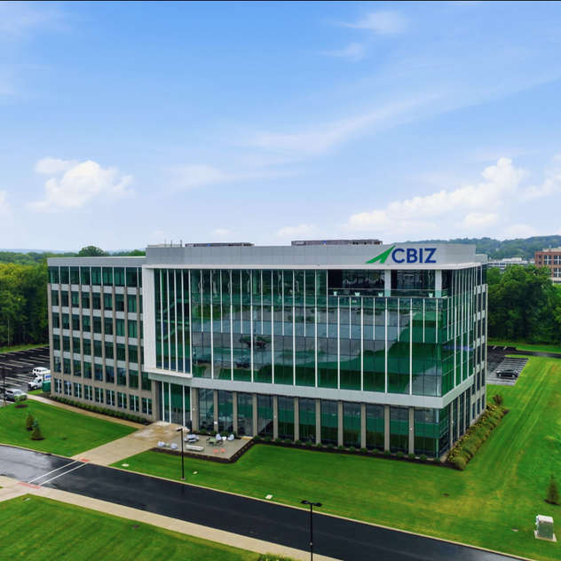 Modern glass office building with green landscaping, cloudy blue sky, and adjacent parking lot.