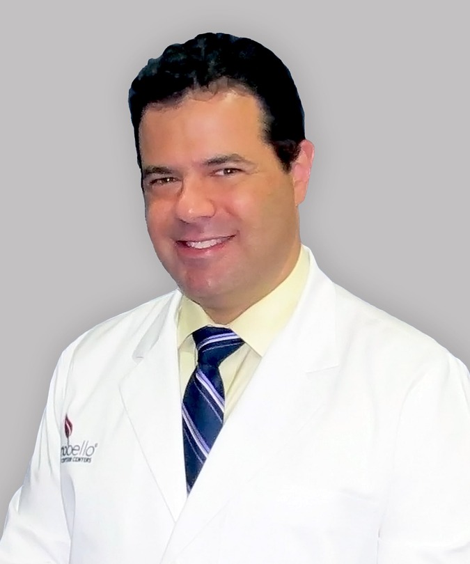 Man in a white lab coat and striped tie smiling against a gray background.