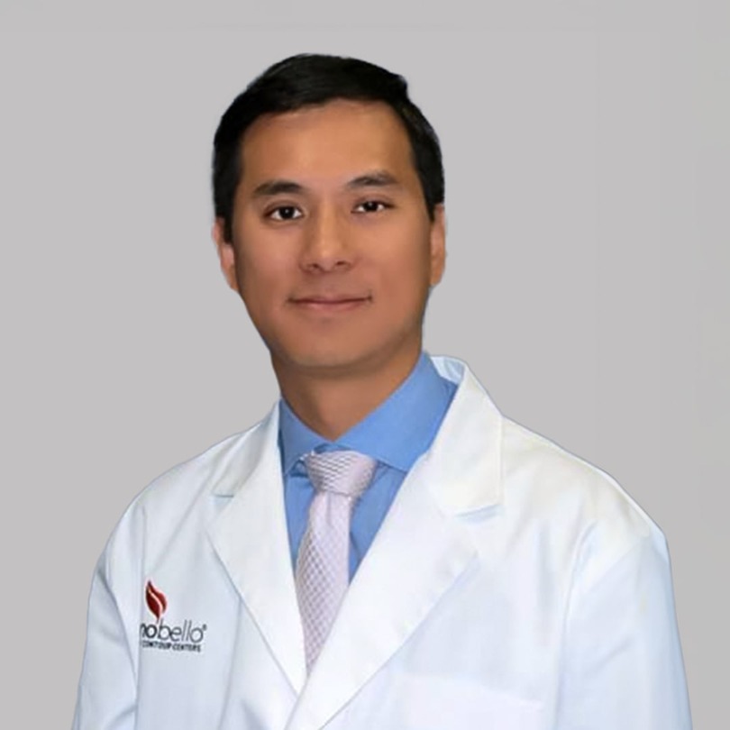 Man in a white coat with a blue shirt and pink tie, standing against a plain background.