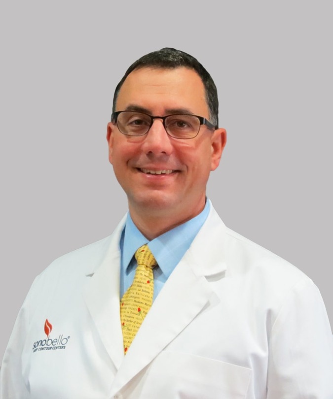 A smiling doctor in a white coat with a yellow tie standing against a gray background.