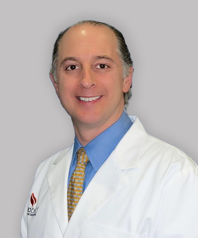 Smiling doctor in a white coat with a blue shirt and yellow tie against a gray background.