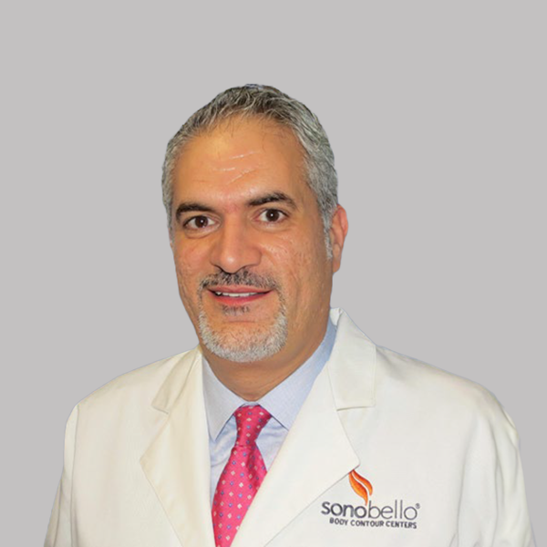 Man in a white medical coat with a company logo, wearing a pink tie and smiling, against a plain gray background.