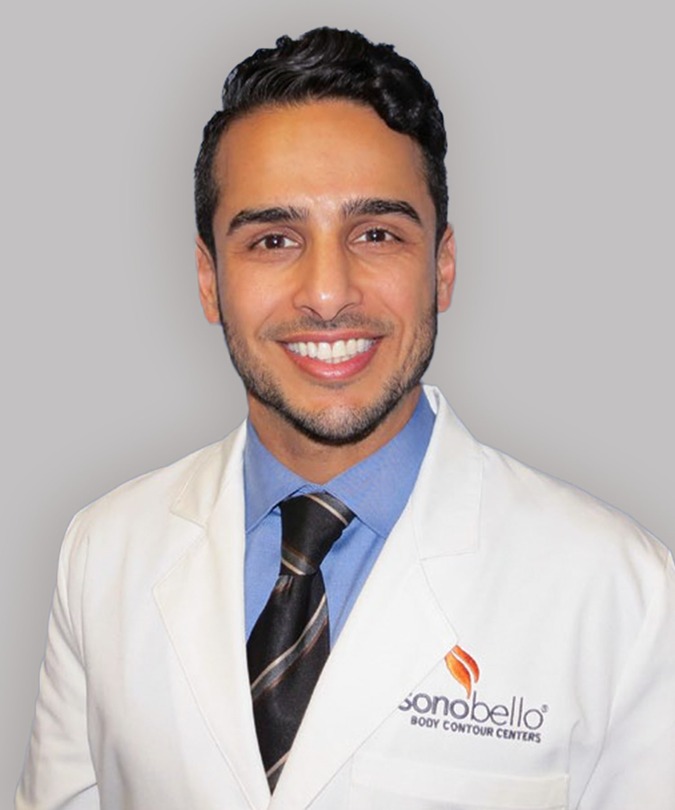 Smiling person in a white coat with a logo on the pocket, standing against a gray background.