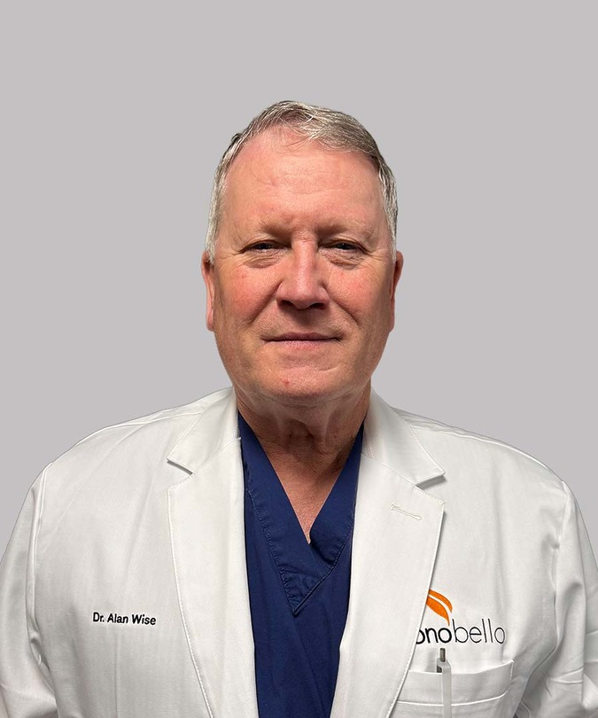A man wearing a white lab coat over blue scrubs, standing against a neutral background.