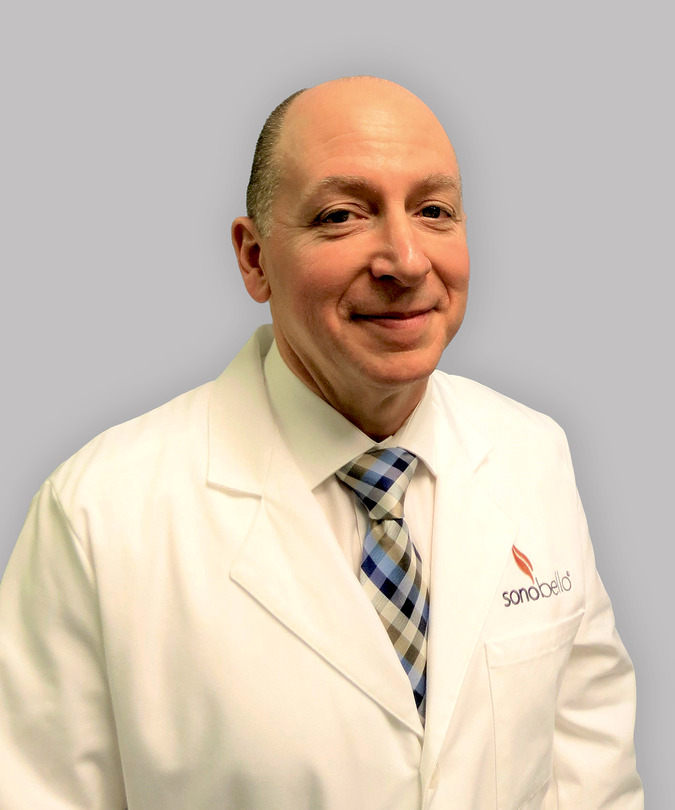 Man in a white coat and plaid tie smiling against a gray background.
