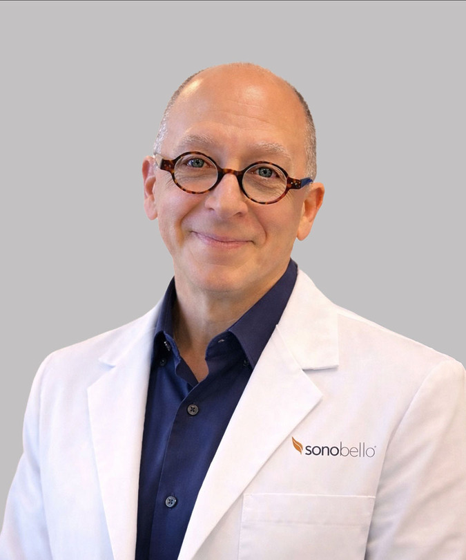 Bald man wearing glasses and a white lab coat with a Sono Bello logo, smiling against a gray background.