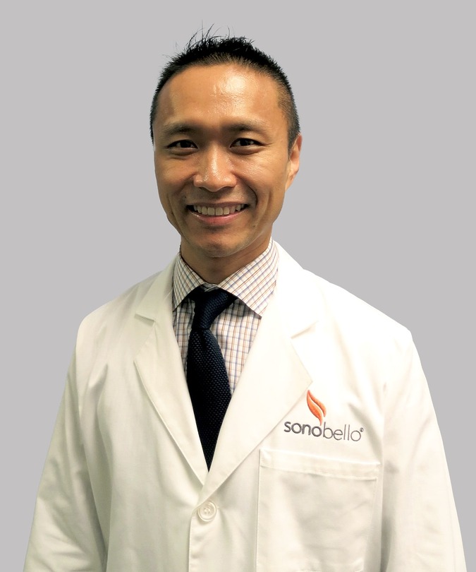 Smiling person wearing a white lab coat with a "Sono Bello" logo, standing in front of a light gray background.