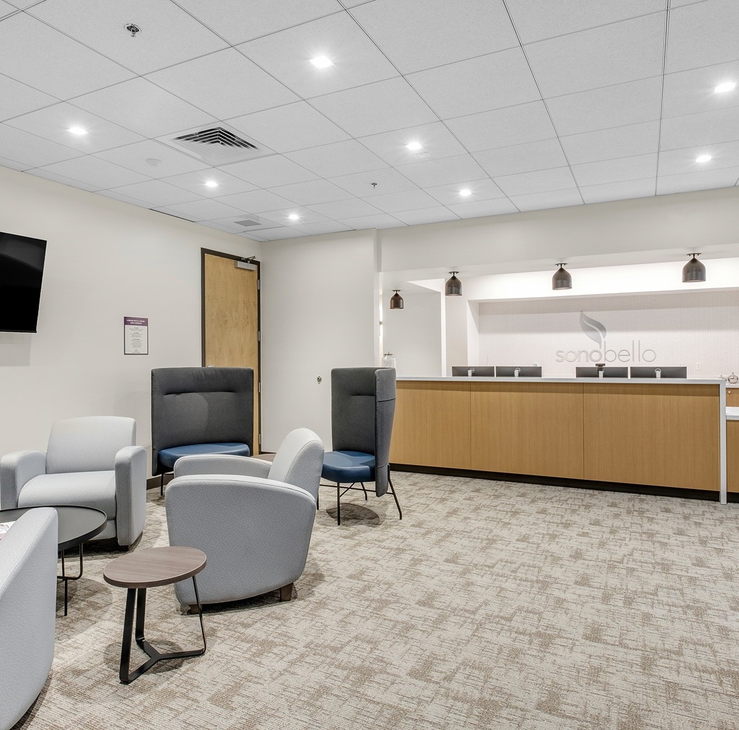 Modern office waiting room with chairs, tables, a wall-mounted TV, and a countertop with overhead lighting.