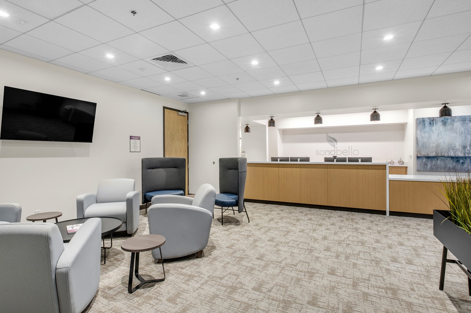 Modern office waiting area with white and gray chairs, a wooden counter, and a wall-mounted TV.