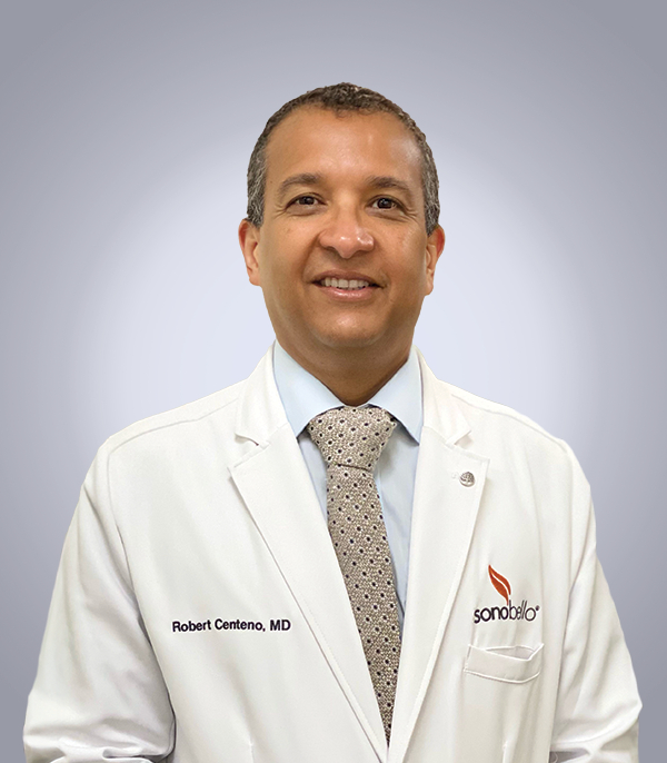 A doctor wearing a white coat with a badge standing against a gray background, smiling at the camera.