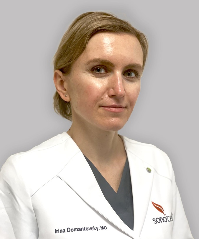 Doctor in a white coat with short blonde hair facing forward against a neutral background