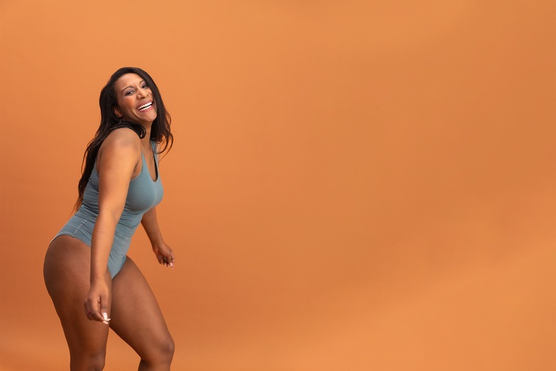 Woman in a blue dress smiling and posing playfully against an orange background.