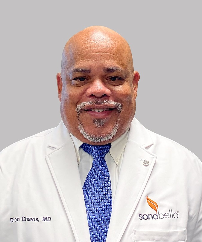 Smiling man in a white doctor's coat with a blue tie, facing forward against a light gray background.