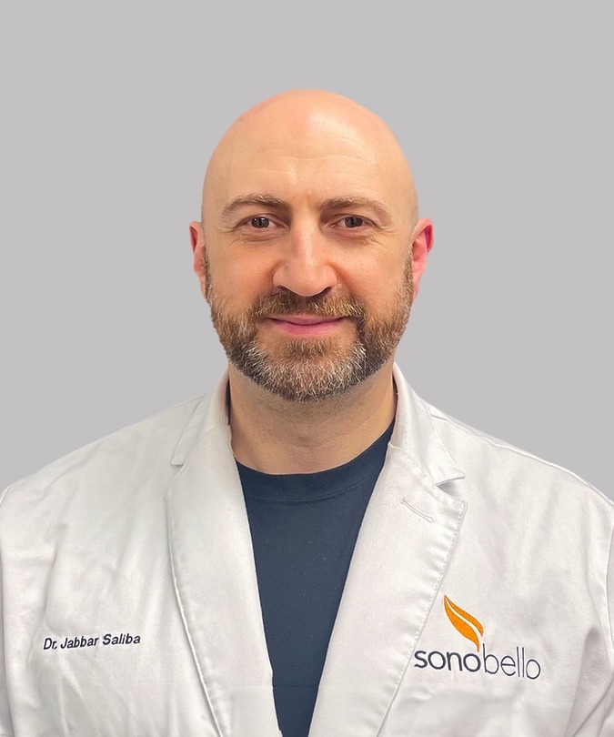 A person wearing a white lab coat with a logo on the chest, standing against a plain background.