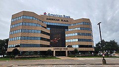 Modern multi-story office building with glass windows, "American Bank" signage, surrounded by trees and a parking lot.