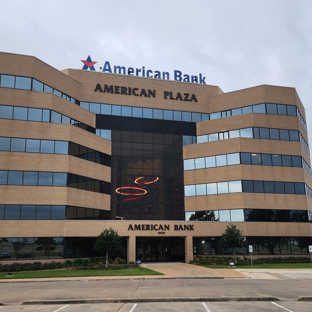 Modern multi-story office building with glass windows, "American Bank" signage, surrounded by trees and a parking lot.