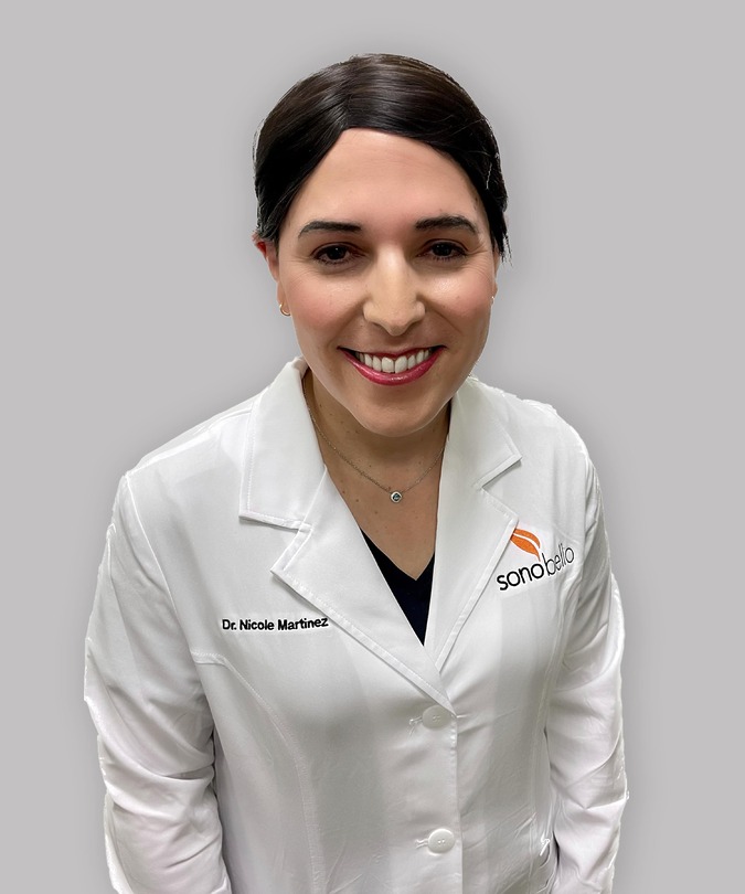 Female doctor smiling in white lab coat with dark hair against a gray background.