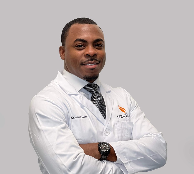 Male doctor in a white coat smiling, arms crossed, wearing a black tie and wristwatch against a light gray background.