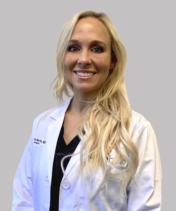 Smiling person in a white lab coat with long blonde hair against a gray background.