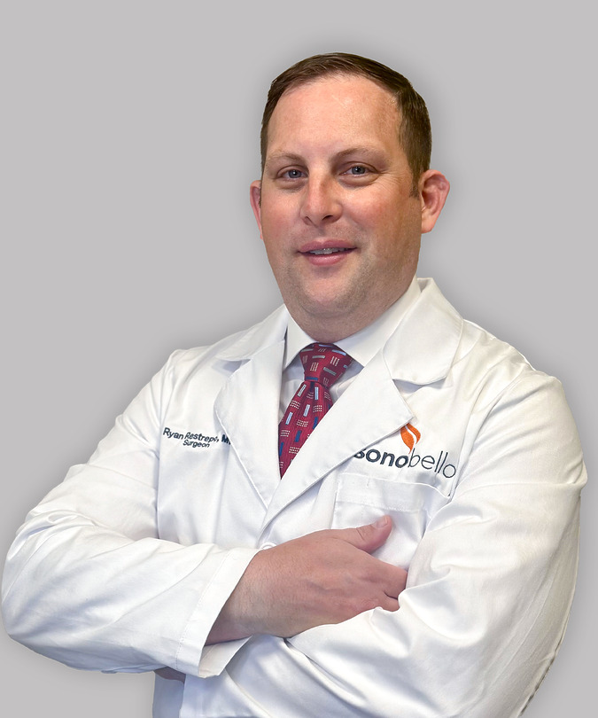 Doctor in a white coat with arms crossed, wearing a red tie, against a plain background.