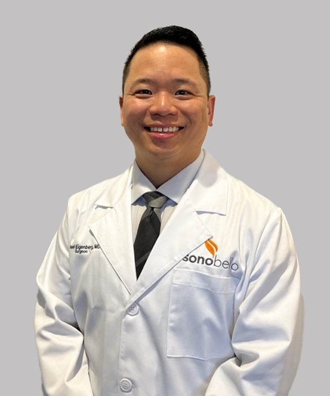 Man in a white lab coat smiling, standing against a gray background