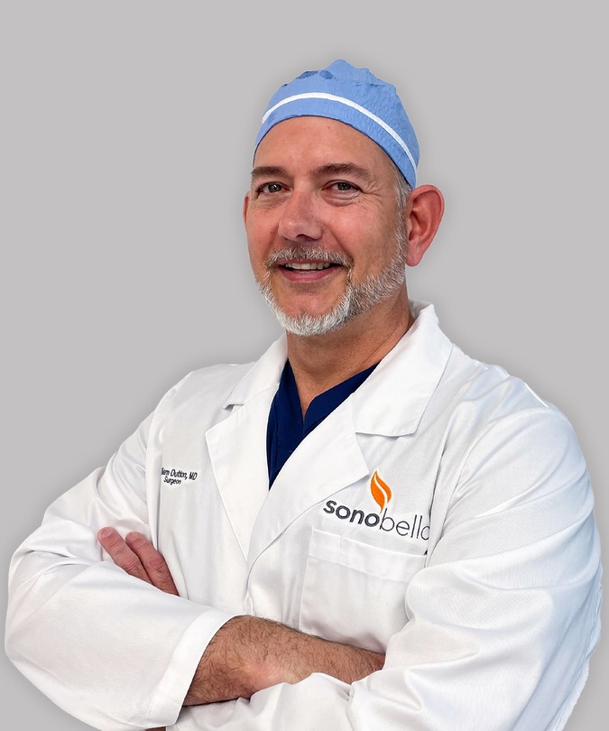 Smiling male doctor in a white coat and blue surgical cap with arms crossed against a gray background.
