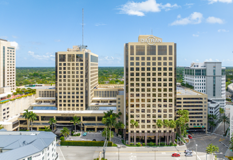 Two tall office buildings with palm trees in front and a clear blue sky in the background.