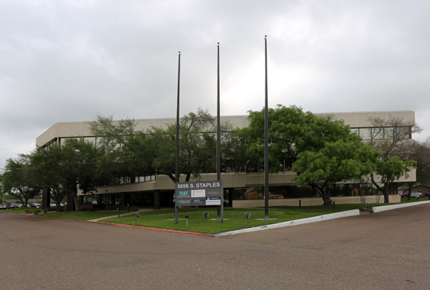 Office building exterior with entrance, surrounding trees, and a sign displaying business names on a cloudy day.