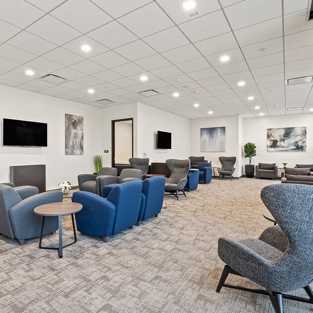Modern office lounge with blue and gray chairs, carpeted floor, and wall-mounted TVs