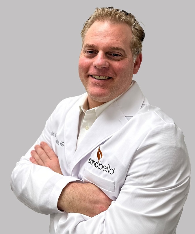 A smiling person in a white lab coat with folded arms against a gray background.