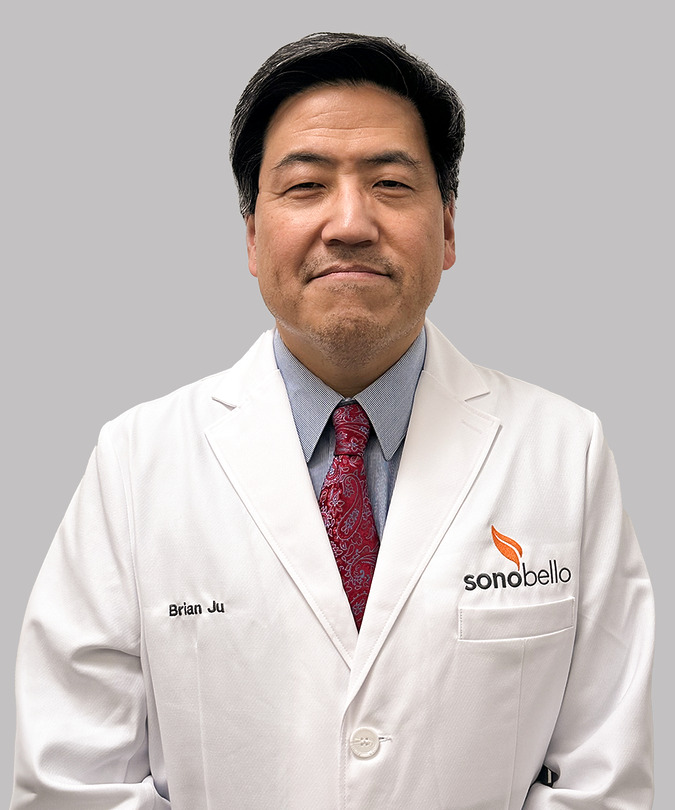Male doctor in a white coat with a red tie, standing against a plain background.