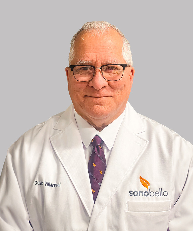 A person wearing glasses and a white lab coat with a logo, standing in front of a light gray background.