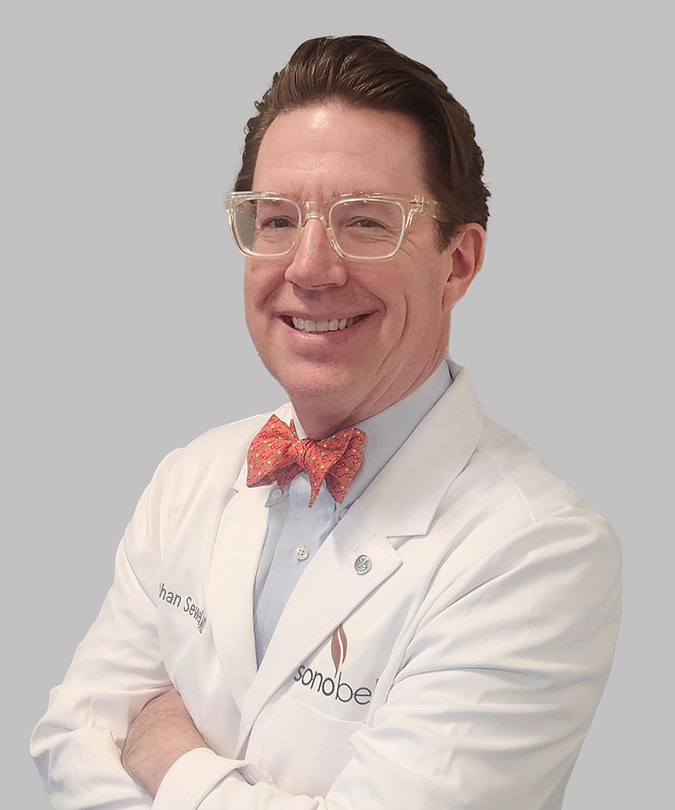 Man in a white lab coat and red bow tie, smiling, with crossed arms against a light gray background.