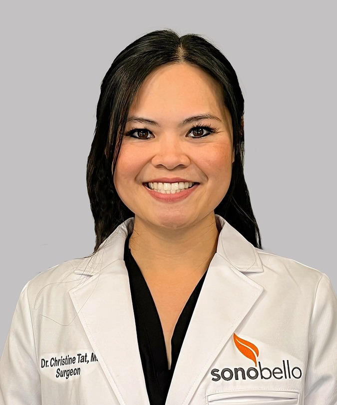 Smiling individual wearing a white coat against a gray background.