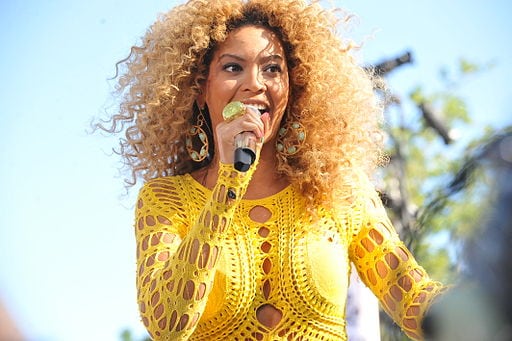 Singer performing on stage, wearing a yellow crochet top and holding a microphone with curly hair.