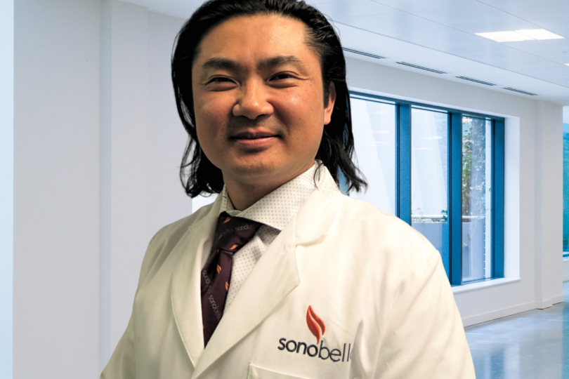 Person in a white lab coat standing in a bright, modern office space with large windows in the background.