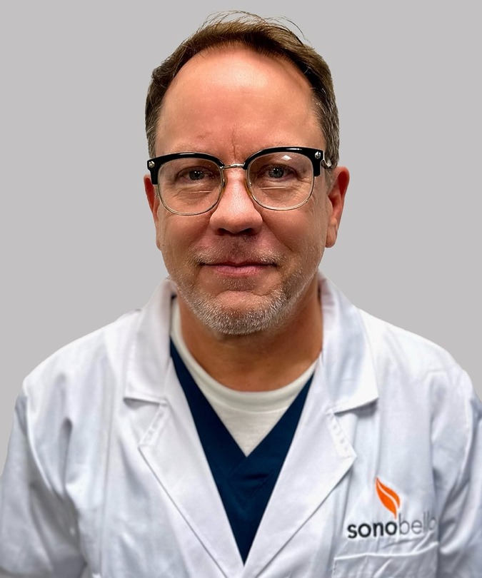 Man wearing glasses and a white lab coat with a logo, standing against a neutral background.