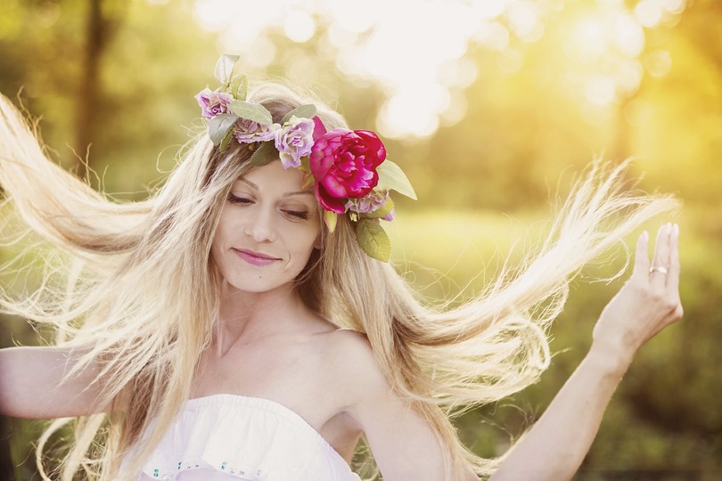 Woman in a white dress with a floral crown of pink and red flowers, standing in a sunlit field, eyes closed, hair flowing.