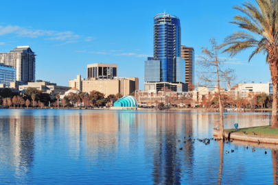 Orlando skyline with modern buildings and palm trees reflected in a calm lake under a clear blue sky.