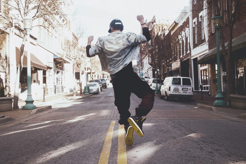 Person jumping joyfully on a city street, wearing a cap and urban attire, with buildings and vehicles in the background.