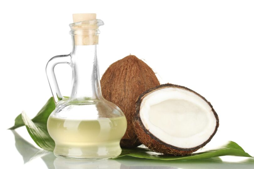 Glass bottle of coconut oil next to whole and halved coconuts on a green leaf background.