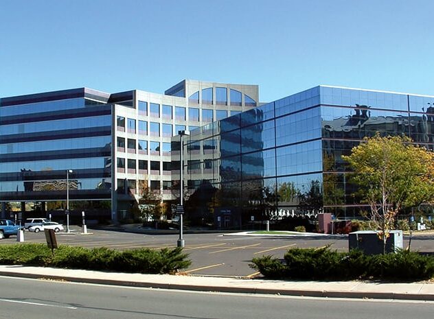 Modern office building with glass facade reflecting the sky and surrounding trees on a clear day.
