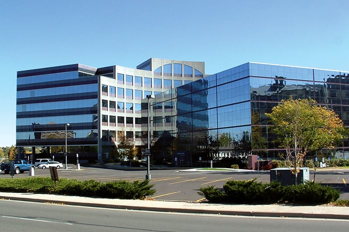 Modern office building with glass facade reflecting the sky and surrounding trees on a clear day.