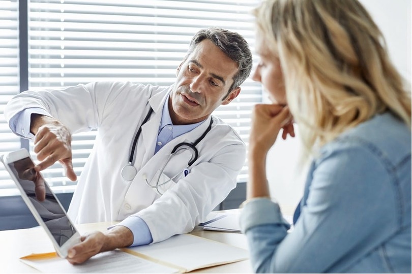 Doctor explaining medical results to a patient seated at a desk, with blinds in the background.