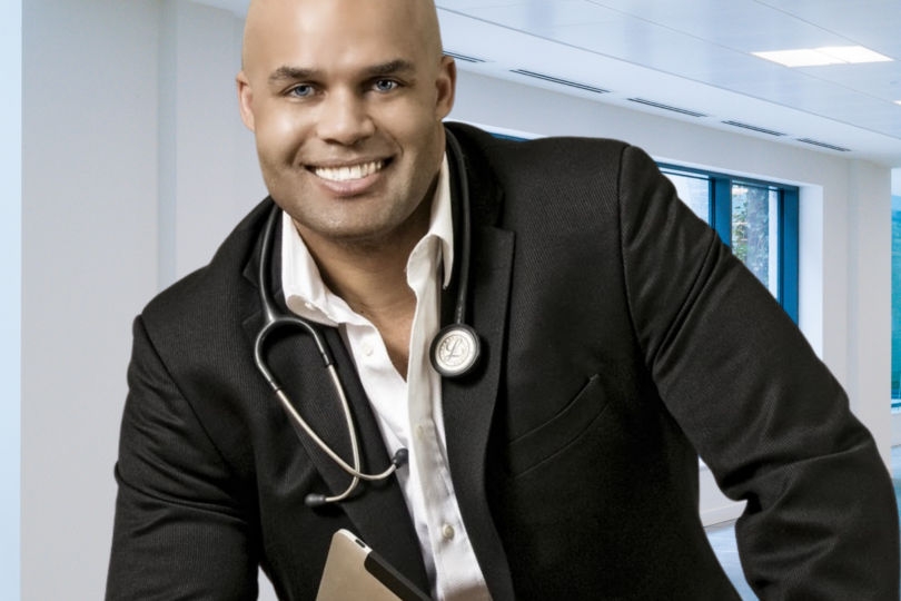 Smiling man in a suit with a stethoscope around his neck, holding a tablet, standing in a modern office setting.