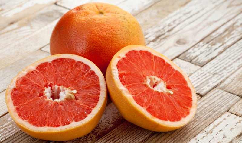 Sliced grapefruit on wooden surface with whole grapefruit in the background.