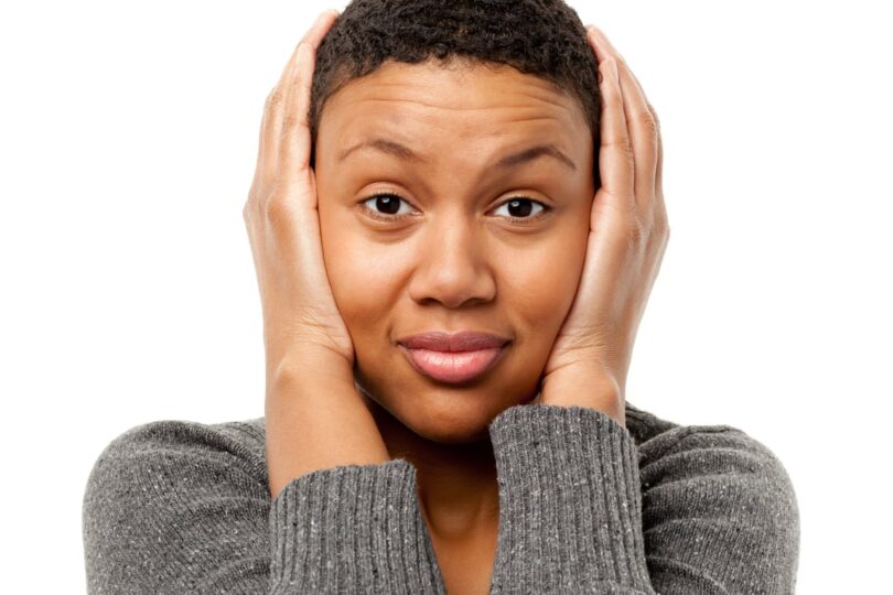 Person covering ears with hands, wearing a grey sweater, looking directly at the camera against a white background.