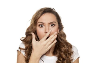 Person with long hair looks surprised, covering mouth with one hand against plain white background.